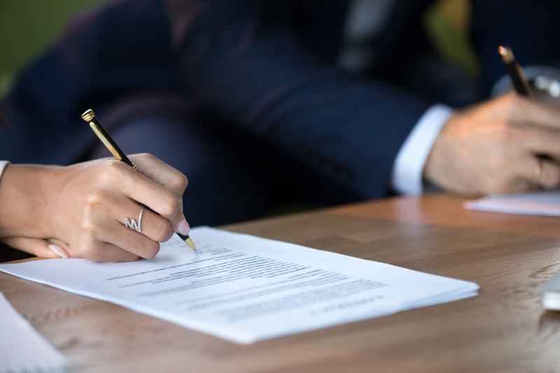 A closeup image of two business partners signing settlement agreement contracts at a desk.